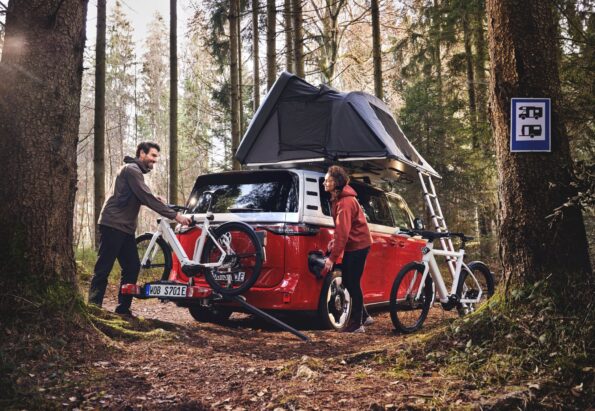 Two people load bicycles onto a roof rack of a red camper van with a raised tent in a pine forest.